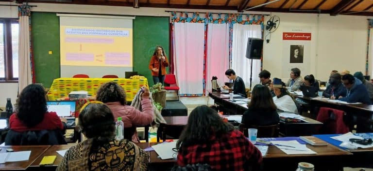 Woman wearing red with long hair and glasses speaks into a microphone in front of an auditorium in Brazil.